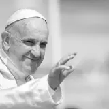 Papst Franziskus winkt lächelnd in die Menge, gekleidet in seine Soutane und mit einer Kette.