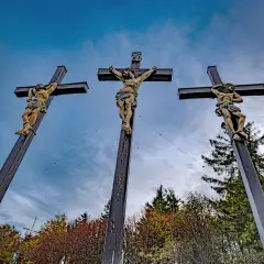 Drei Kreuze mit Jesusfiguren, die die Passion Christi darstellen, vor einem dramatischen Himmel und herbstlichem Laub.