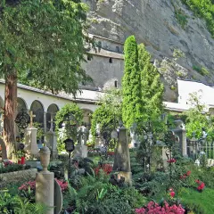 Gräberfeld mit Kreuzen und Urnen vor Arkaden der Erzabtei Salzburg. Üppige Vegetation und Felswand im Hintergrund.