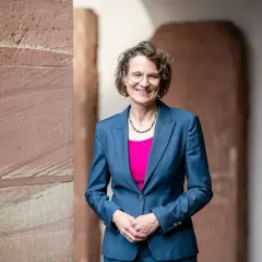 Christiane Tietz, eine Frau mit Brille und lockigem Haar, lächelt in einem blauen Hosenanzug vor einer Sandsteinwand.