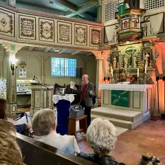 Ein Mann spricht in einer Kirche vor Zuhörern. Er hält ein Buch hoch, das von seelischem Widerstand handeln könnte.