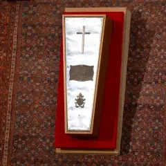 Der gläserne Sarg eines Papstes, mit Kreuz und Wappen, liegt auf einem roten Samtkissen vor einem reich gemusterten Teppich im Petersdom.