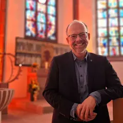 Lächelnder Mann in Anzug lehnt an einer Kirchenbank. Buntglasfenster und Taufbecken im Hintergrund. Hier kann man wieder in die Kirche eintreten.