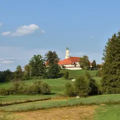 Kloster Reutberg thront auf einem Hügel, umgeben von grünen Wiesen und Bäumen unter blauem Himmel mit Wolken.