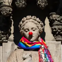 Steinskulptur mit roter Clownsnase und buntem Schal. Die Frage "was hat Karneval mit Religion zu tun?" wird hier humorvoll aufgeworfen.