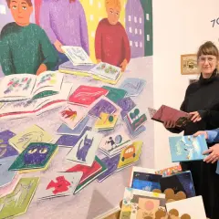 Zwei Frauen stehen vor einer bunten Wandmalerei mit Büchern und halten Bücher in den Händen.