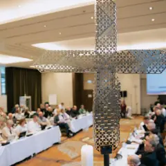 Ein moderner Kreuz-Schriftzug dominiert die Szene, während im Hintergrund ein synodaler Ausschuss tagt.