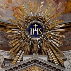 Goldene Strahlenkrone mit IHS-Monogramm, das die Bedeutung von Jesus Christus symbolisiert, umgeben von Engeln und Putten.