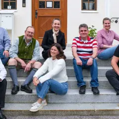 Eine Gruppe von Menschen sitzt lächelnd auf einer Treppe. Sie scheinen Teil einer Gemeinschaft zu sein, vielleicht ein Dekanat, das sich für ein Foto versammelt hat.