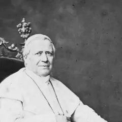 Papst Pius sitzt auf einem prunkvollen Thron, gekleidet in seine Soutane. Neben ihm steht ein aufwendig verziertes Kreuz.