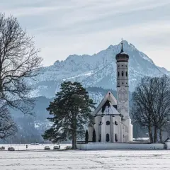 Winterliche Szene mit der Kirche St. Coloman im bayerischen Voralpenland, schneebedeckte Berge im Hintergrund.