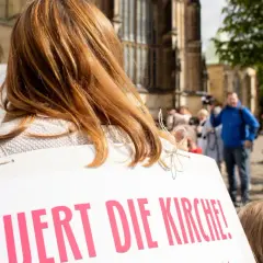 Frauen mit Schildern fordern "Erneuert die Kirche!". Eine Frau mit roten Haaren und eine mit blonden Haaren sind zu sehen.