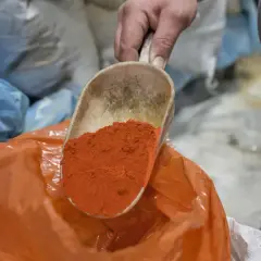 Eine Schaufel mit leuchtend orangefarbenem Pulver wird über einen orangefarbenen Beutel gehalten. Die Farben und ihre Bedeutung sind Teil eines alten Handwerks.