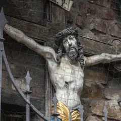 Ein detailreiches, altes jesus kreuz original, hinter einem Metallgitter und Netz, zeigt die leidende Figur Christi vor einer Steinwand.