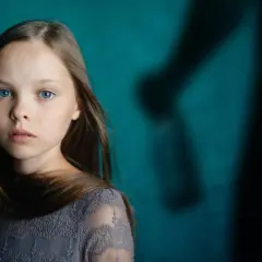 Ein junges Mädchen mit blauen Augen blickt nachdenklich, während im Hintergrund eine schattenhafte Figur erscheint, die an einen beschützenden Gott als Vater erinnert.