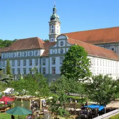 Prächtiges Kloster FFB mit barockem Turm, umgeben von grünen Bäumen und einem lebhaften Markt im Vordergrund.
