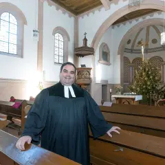 Ein Geistlicher in der Kirche, der die Bänke zeigt. Er wirkt freundlich, als würde er die Bedeutung von Gemeinschaft und Glauben erklären, im Gegensatz zur simonie definition.