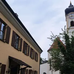 Ein Haus mit braunen Fensterläden und ein Kirchturm mit Uhr sind im Pfarrverband Erdinger Moos zu sehen.