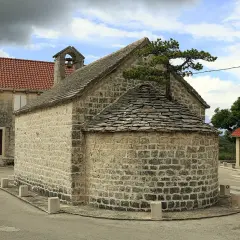 Kleine Steinkirche mit halbrunder Altarnische, bedeckt mit Schindeln. Ein Baum wächst aus dem Dach.