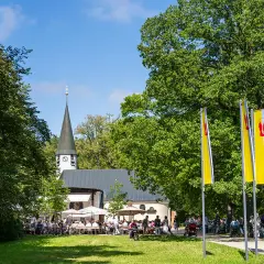Ein sonniger Tag bei Maria Eich Planegg. Menschen sitzen unter Sonnenschirmen vor der Kirche, während Fahnen im Wind wehen.