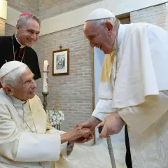 Papst Franziskus und ein Bischof lächeln, während sie Benedikt XVI. die Hand schütteln.