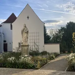 Statue vor dem Domberg Freising. Ein Weg führt durch einen blühenden Garten zu einem Tor.
