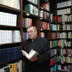 Kardinal Lehmann steht in einer Bibliothek und hält ein Buch in den Händen. Um ihn herum sind Regale voller Bücher.