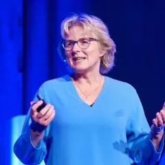 Melanie Wolfers, eine inspirierende Rednerin, hält einen Vortrag vor einem blauen Hintergrund. Sie trägt eine blaue Bluse und eine Halskette.