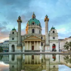 Die Karlskirche Wien spiegelt sich im Wasser. Majestätische Säulen flankieren die Kuppel. Ein dramatischer Himmel über der barocken Pracht.