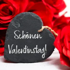 Herzförmiges Schild mit der Aufschrift "Schönen Valentinstag!" vor roten Rosen. Ein liebevoller Gruß zum heiligen Valentin.
