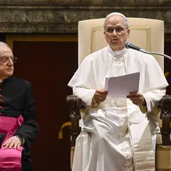 Papst Franziskus spricht in ein Mikrofon, während ein Kardinal daneben sitzt.
