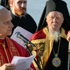 Zwei Geistliche, einer im roten Gewand, der andere mit Bart und Mitra, stehen vor Mikrofonen. Der Mann mit Mitra ist ein Patriarch.