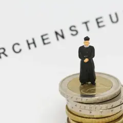 Miniatur-Priester steht auf Münzstapel. Der Schriftzug "Kirchensteuer" deutet auf die Verwendung der Gelder hin.