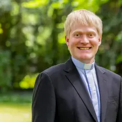 Thomas Frings, ein Geistlicher mit blondem Haar, lächelt freundlich in die Kamera. Er trägt einen schwarzen Anzug mit einem blauen Hemd und einem weißen Kragen.