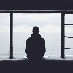 Eine Person blickt aus einem Fenster auf das Meer. Schnee liegt auf dem Sims. Das Bild vermittelt ein Gefühl von Einsamkeit.