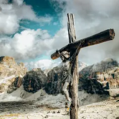 Holzfigur der Jesus-Kreuzigung vor dramatischer Bergkulisse und Wolkenhimmel.
