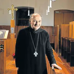 Ein lächelnder Odilo Lechner in schwarzer Soutane mit Kreuzkette steht in einer Kirche vor Beichtstühlen und einem Lesepult.