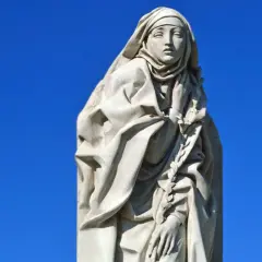 Steinskulptur der Heiligen Katharina von Siena mit Schleier und einem Lorbeerkranz vor blauem Himmel.