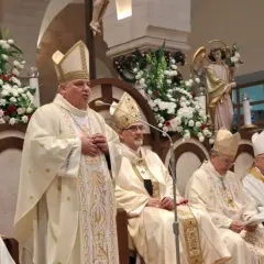 Bischof Konrad Krajewski spricht zu einer Gruppe von Geistlichen in festlicher Kleidung, umgeben von Blumen und religiösen Statuen.