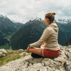 Frau meditiert auf einem Felsen mit Blick auf ein alpines Tal. Sie findet Ruhe und Inspiration für ihre Meditationstexte unterwegs sein.