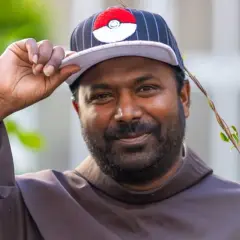 Pater Sandesh Manuel, ein Mann mit Bart, trägt eine Kappe mit Pokémon-Motiv und eine braune Robe. Er lächelt und hebt die Kappe an.