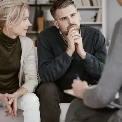Ein Paar sitzt auf einer Couch und spricht mit einer Person, die Notizen macht. Sie sind in einer Sitzung zur Eheberatung.