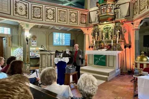 Ein Mann spricht in einer Kirche vor Zuhörern. Er hält ein Buch hoch, das von seelischem Widerstand handeln könnte.
