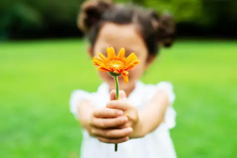 Ein Kind reicht eine orange Blume. Das Gegenteil von Habgier ist Großzügigkeit.