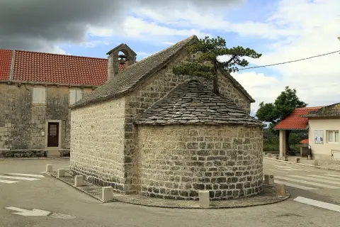 Kleine Steinkirche mit halbrunder Altarnische, bedeckt mit Schindeln. Ein Baum wächst aus dem Dach.