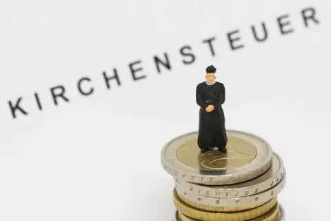 Miniatur-Priester steht auf Münzstapel. Der Schriftzug "Kirchensteuer" deutet auf die Verwendung der Gelder hin.