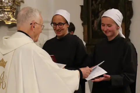 Priester übergibt Dokumente an zwei Dienerinnen vom Heiligen Blut, die lächelnd ihre Aufgaben annehmen.