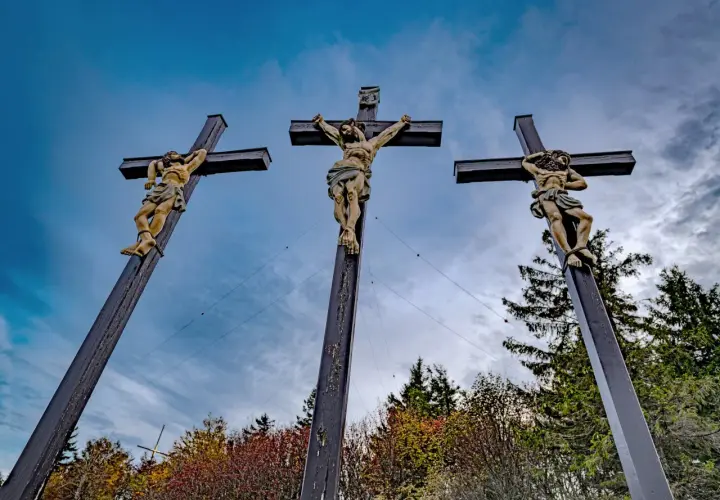 Drei Kreuze mit Jesusfiguren, die die Passion Christi darstellen, vor einem dramatischen Himmel und herbstlichem Laub.