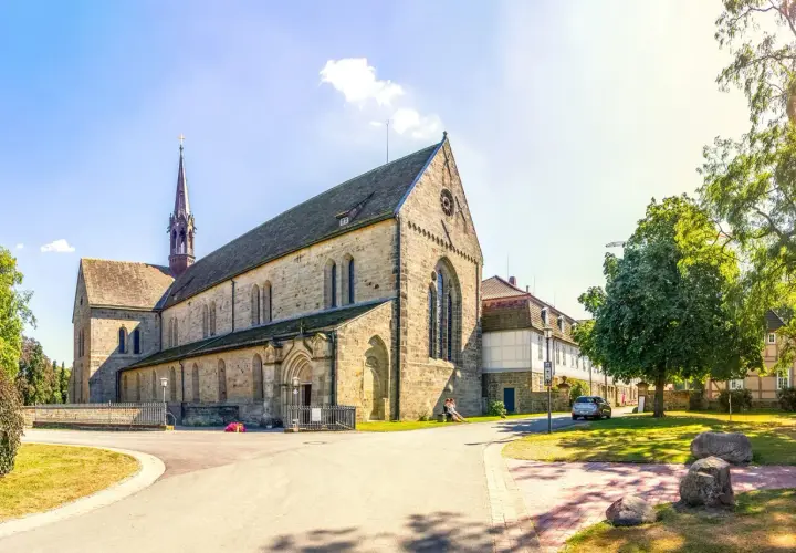 Klosterkirche mit Turm, Teil des Jakobsweg Hessen. Ein Paar sitzt auf einer Bank im Schatten eines Baumes.