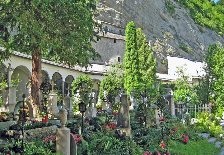 Gräberfeld mit Kreuzen und Urnen vor Arkaden der Erzabtei Salzburg. Üppige Vegetation und Felswand im Hintergrund.